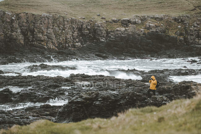 Viajante masculino em pé na rocha com câmera no tripé e tirar fotos da paisagem marinha no dia sombrio nublado na costa da Irlanda do Norte — Fotografia de Stock