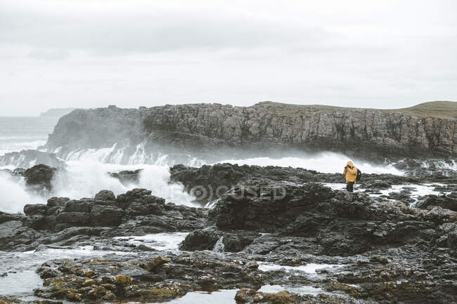 Hombre viajero de pie en la roca con la cámara en el trípode y tomar una foto del paisaje marino en el día nublado y sombrío en la costa de Irlanda del Norte - foto de stock