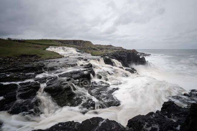 Sea waves crashing on rocks and breaking down to splashes on stormy day with heavy clouds at Northern Ireland coastline — Stock Photo
