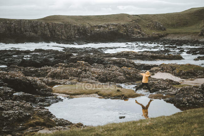 Vista lateral del turista saltando sobre charcos dejados con agua de mar en la costa rocosa mientras camina en la costa de Irlanda del Norte en el nublado día de primavera - foto de stock