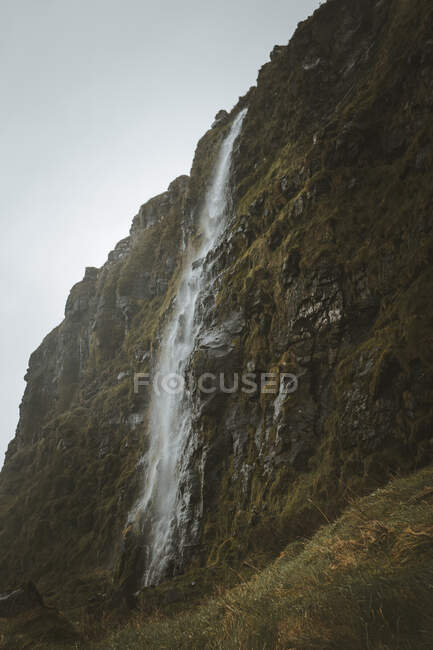 Green meadow at foot of cliff with waterfall against grey cloudy sky in spring day in Northern Ireland — Stock Photo