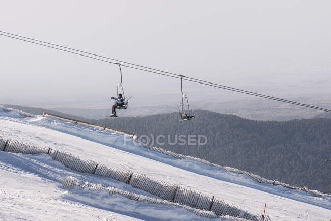 Vista lateral da pessoa irreconhecível com snowboard montando teleférico sobre encosta de montanha nevada no dia de inverno no resort — Fotografia de Stock