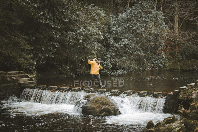 Seitenansicht eines männlichen Touristen in leuchtend orangefarbener Jacke, der auf einer Fußgängerbrücke läuft und den Fluss überquert, wobei Wasser durch Trittsteine im Wald von Nordirland fließt — Stockfoto
