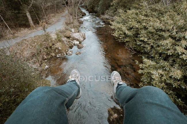 Вище врожай мандрівник у джинсах і кросівках сидить на краю моста і звисає ногами по річці під час прогулянок у весняному лісовому парку Північної Ірландії. — стокове фото