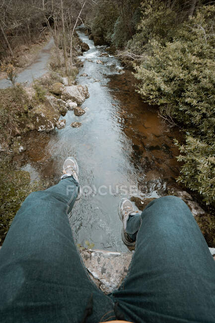 De cima viajante colheita em jeans e tênis sentado na borda da ponte e balançando as pernas sobre o rio durante caminhadas no parque florestal primavera na Irlanda do Norte — Fotografia de Stock