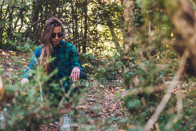 Long haired female hipster in stylish casual plaid shirt sneakers and trendy sunglasses sitting on small stub on spangled of leafage path among green trees — Stock Photo
