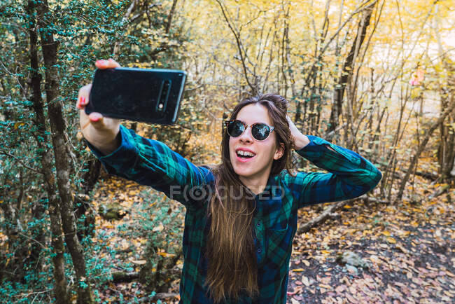 Woman in plaid shirt taking selfie on mobile phone while standing on forest wood — Stock Photo