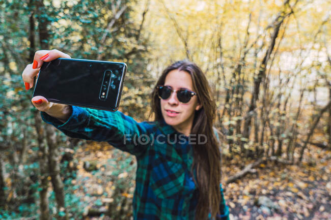 Frau im karierten Hemd macht Selfie mit Handy, während sie auf Waldholz steht — Stockfoto