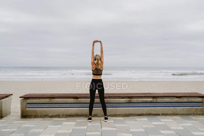 Зворотній вигляд спортсменки в активному носінні, що стоїть з піднятими руками і тягнеться біля берега моря на похмуру погоду — стокове фото