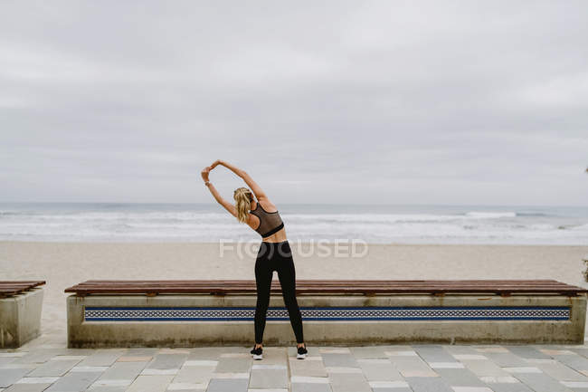 Зворотній вигляд спортсменки в активному носінні, що стоїть з піднятими руками і тягнеться біля берега моря на похмуру погоду — стокове фото