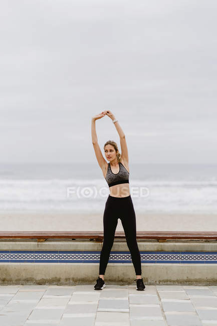 Female athlete in active wear standing with raised hands and stretching near seashore on cloudy weather — Stock Photo