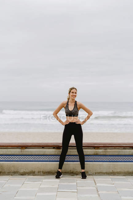Sportlerin in aktiver Kleidung steht bei trübem Wetter mit den Händen auf Hüften in Küstennähe — Stockfoto