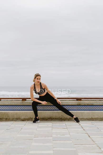 Mujer deportiva motivada en desgaste activo de pie estirando las piernas mientras mira hacia otro lado - foto de stock