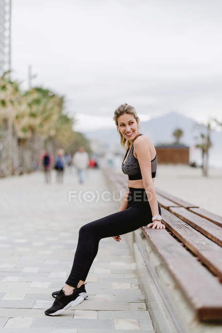 Vista laterale di atleta concentrato in abbigliamento attivo alla moda seduto sulla panchina, guardando lontano con il mare su sfondo sfocato — Foto stock