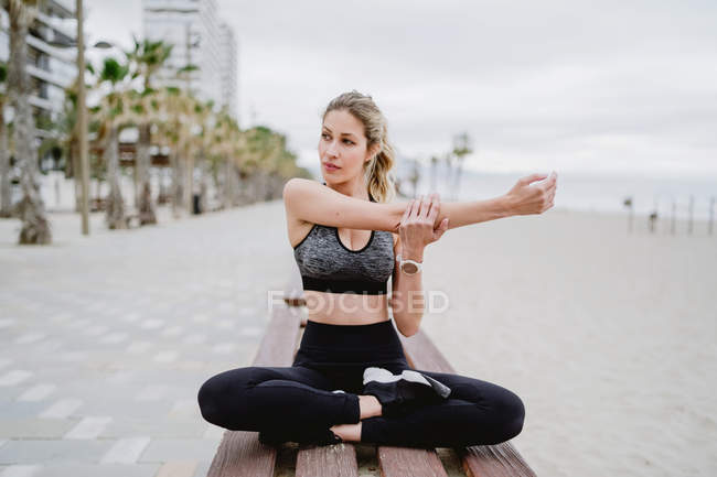 Concentrated cross-legged athlete sitting on bench and stretching arms with seaside on blurred background — Stock Photo