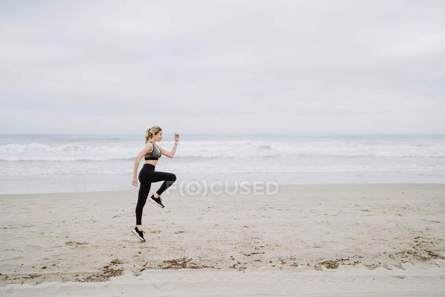 Vista lateral de determinada jogger feminino em roupas esportivas elegantes e tênis pulando na praia vazia em tempo nublado — Fotografia de Stock