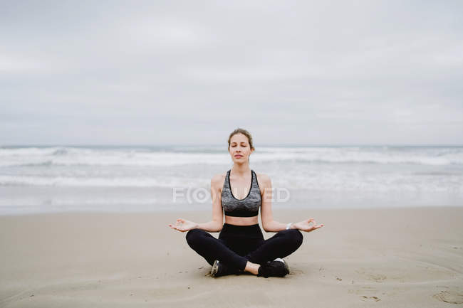 Молода струнка жінка в чорному верху і легінси, сидячи в положенні лотоса на пляжі з закритими очима під час медитації — стокове фото