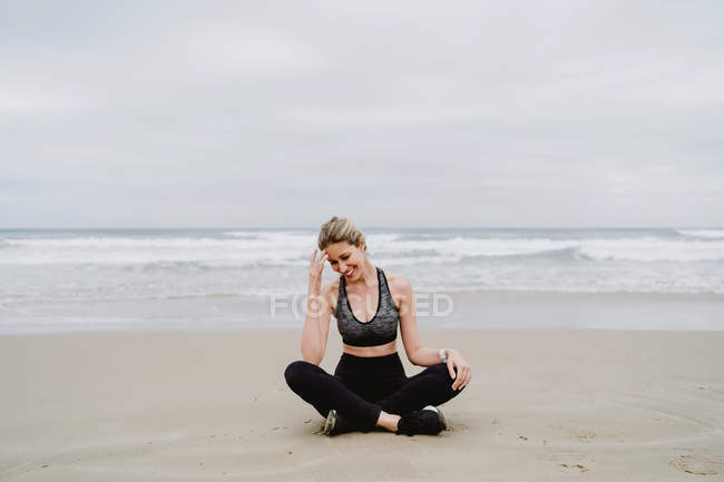 Young slim female in black top and leggings sitting in lotus position and smiling at beach — Stock Photo