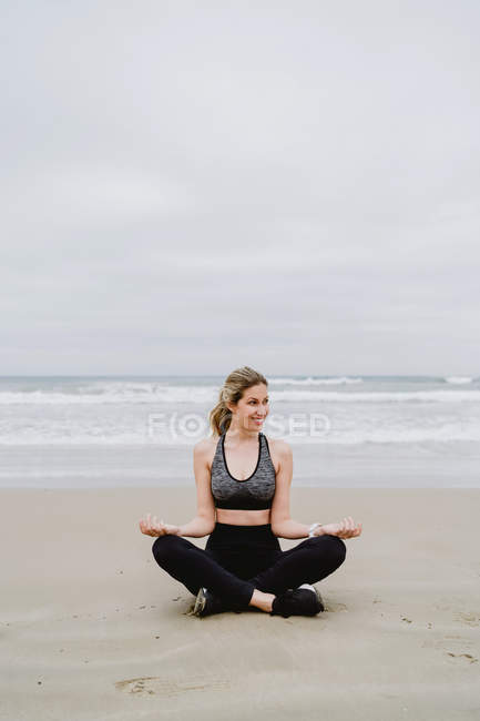 Young slim female in black top and leggings sitting in lotus position and looking away at beach — Stock Photo