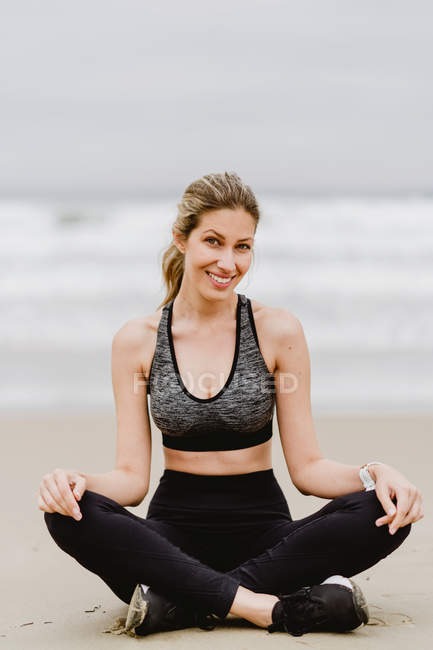 Молодая стройная женщина в черном топе и леггинсы сидят в позе лотоса во время медитации на пляже — стоковое фото