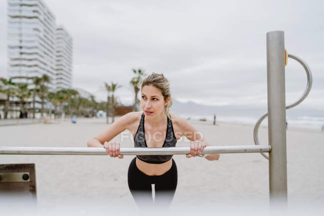 Мотивированная спортивная женщина в активном износе тренируется в металлическом баре на песчаном пляже — стоковое фото