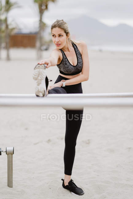 Motivierte sportliche Frau in aktiver Kleidung Stretching in Metallstange am Sandstrand — Stockfoto