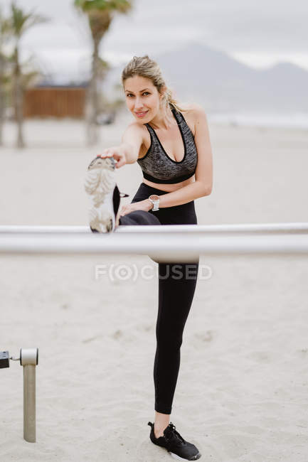 Mujer deportiva motivada en desgaste activo estiramiento en barra de metal en la playa de arena - foto de stock