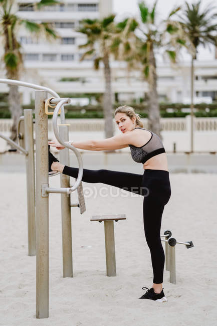 Vista lateral da mulher esportiva motivada no desgaste ativo que se estende no bar de metal na praia arenosa — Fotografia de Stock
