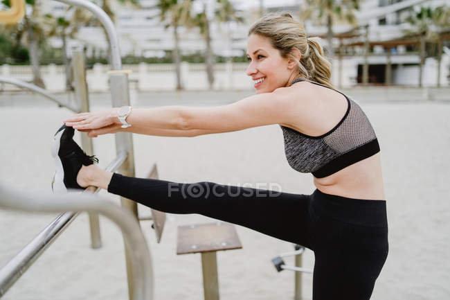 Vue latérale de la femme sportive motivée en tenue active s'étirant dans une barre métallique à la plage de sable — Photo de stock