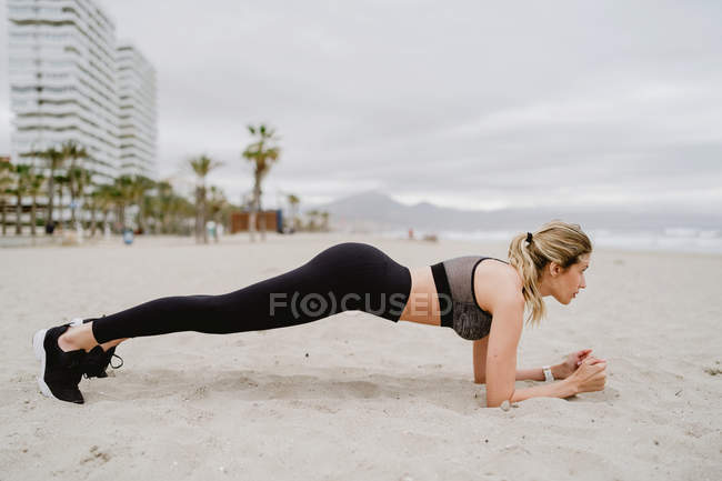 Vista lateral de atleta feminina concentrada em elegante desgaste ativo e tênis fazendo prancha na praia vazia — Fotografia de Stock