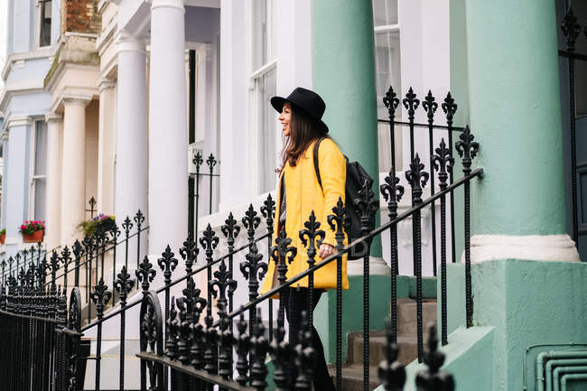 Щаслива стильна жінка в жовтому пальто і капелюсі посміхається, стоячи на сходах у будинку на вулицях Лондона, Велика Британія. — стокове фото