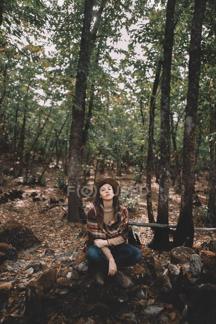 Mujer joven calma en sombrero elegante y bufanda sentado en el suelo con los ojos cerrados y descansando en el bosque verde - foto de stock