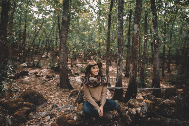 Спокійна щаслива молода жінка в стильному капелюсі і шарфі сидить на землі, дивлячись далеко, відпочиваючи в зеленому лісі — стокове фото
