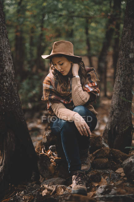 Calm young female in stylish hat and scarf sitting on ground with closed eyes and resting in green forest — Stock Photo