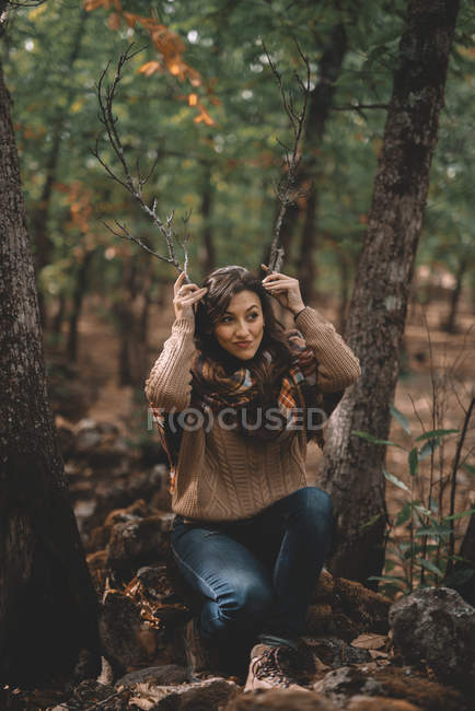Lustige Frau in lässigem Outfit, die Stöcke wie Geweihe in der Hand hält und wegschaut, während sie auf einem Stein im Wald sitzt — Stockfoto