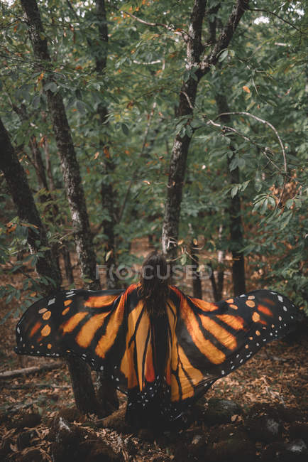 Indietro vista di anonima giovane donna in ali di farfalla mantello ballare vicino agli alberi nella foresta verde — Foto stock