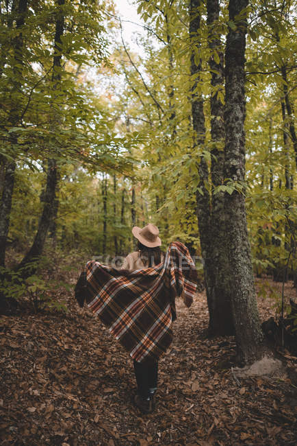Back view of unrecognizable young woman in hat wrapping in checkered scarf while standing on dry leaves in autumn forest — Stock Photo