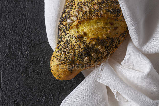 Loaf of ruddy crispy fresh bread with grains and poppy seeds wrapped in towel on grey background — Stock Photo