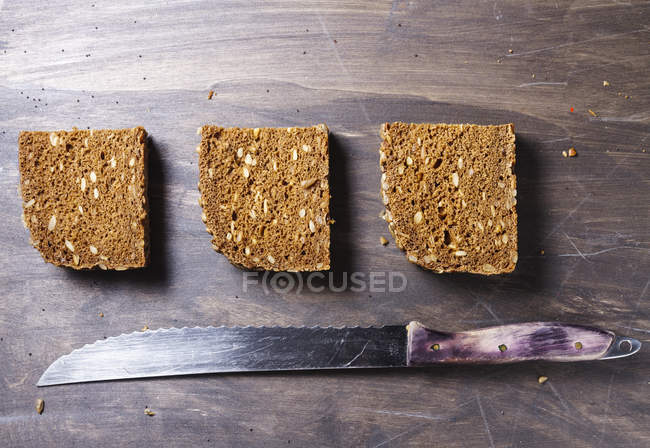 Вид сверху нарезанного коричневого хлеба с целыми булочками и кухонным ножом на деревянном фоне — стоковое фото