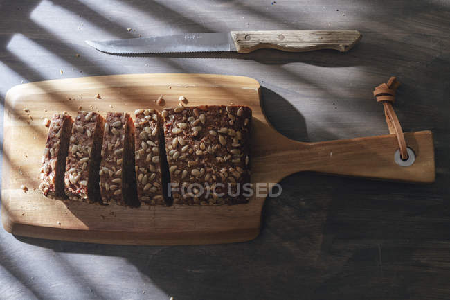 Sliced loaf of wholegrain bread on wooden cutting board and kitchen knife placed on wooden table — Stock Photo