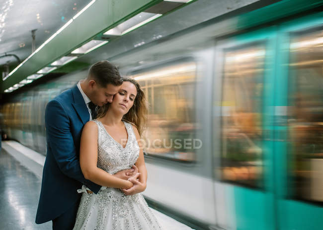 Young bride in white wedding dress standing with closed eyes while groom standing behind and hugging with moving train on blurred background at subway — Stock Photo