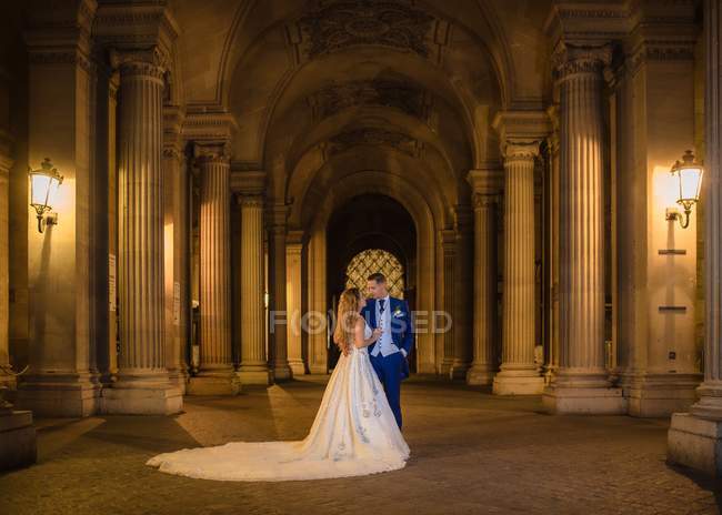 Jovem casal recém-casado em terno de casamento e vestido abraçando enquanto estava em pé em arco balançado com Louvre no fundo em Paris — Fotografia de Stock