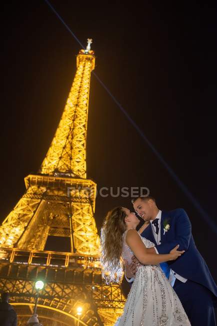 Радостный жених в синем костюме и невеста в белом свадебном платье обнимаются, улыбаясь и целуясь вечером с Эйфелевой башней на заднем плане в Париже — стоковое фото