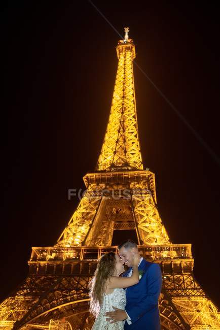 Joyful groom in blue suit and bride in white wedding dress embracing while smiling and kissing on evening with Eiffel Tower on background at Paris — Stock Photo