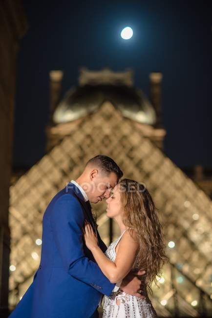 Молода новоодружена пара в весільному костюмі та обнімання під час стоянки в арці з Лувром на задньому плані в Парижі. — стокове фото