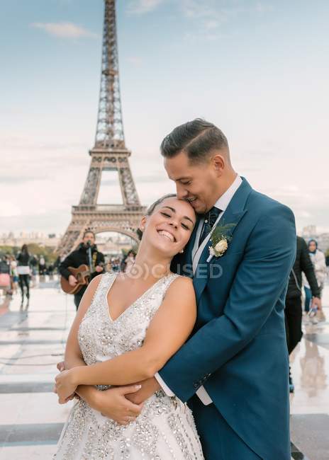 Groom in blue suit and bride in white wedding gown cuddling and smiling with Eiffel Tower on background at Paris — Stock Photo