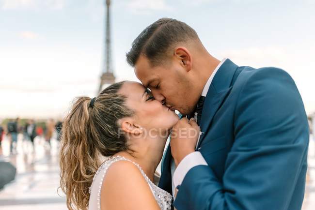 Groom in blue suit and bride in white wedding gown kissing passionately with Eiffel Tower on background at Paris — Stock Photo
