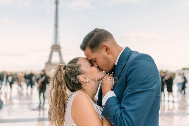 Novio en traje azul y novia en vestido de novia blanco besándose apasionadamente con la Torre Eiffel en el fondo en París - foto de stock
