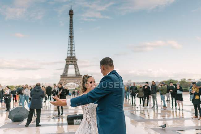 Novio en traje azul y novia en vestido de novia blanco teniendo baile lento sonriendo y mirándose el uno al otro con la Torre Eiffel en el fondo en París - foto de stock