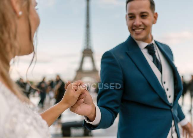 Bräutigam im blauen Anzug und Braut im weißen Hochzeitskleid mit langsamem Tanz lächelnd und einander mit Eiffelturm auf Hintergrund in Paris anschauend — Stockfoto